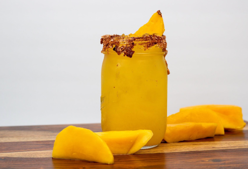 A jar of yellow drink with a sugared rim, garnished with a mango slice, surrounded by fresh mango pieces on a wooden surface.